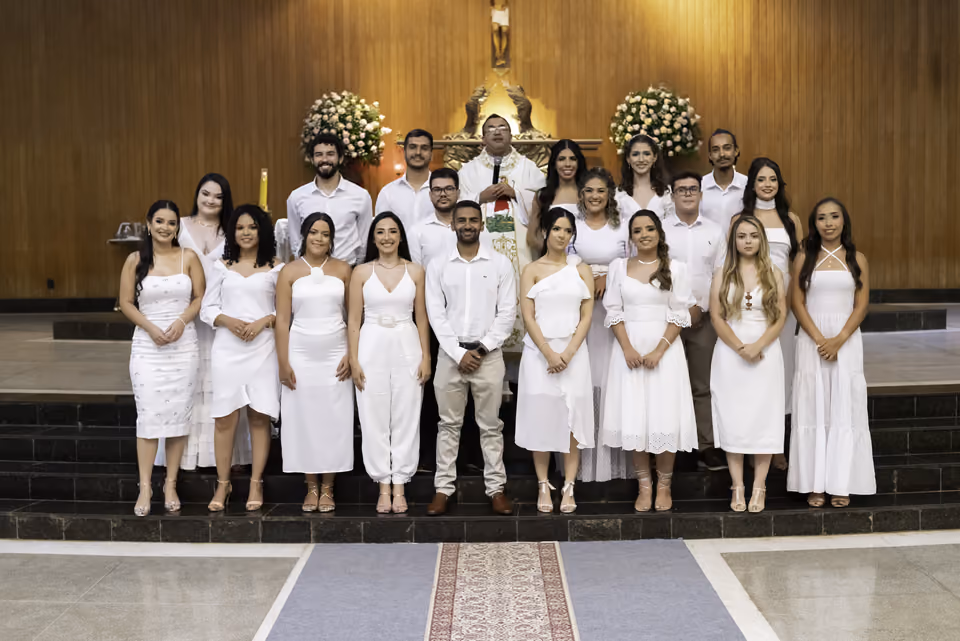 Turma de formandos vestida de branco posa com o celebrante diante do altar, ao fim da missa de formatura.
